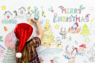 une enfant avec un bonnet de père noël dessine des dessins de Noël